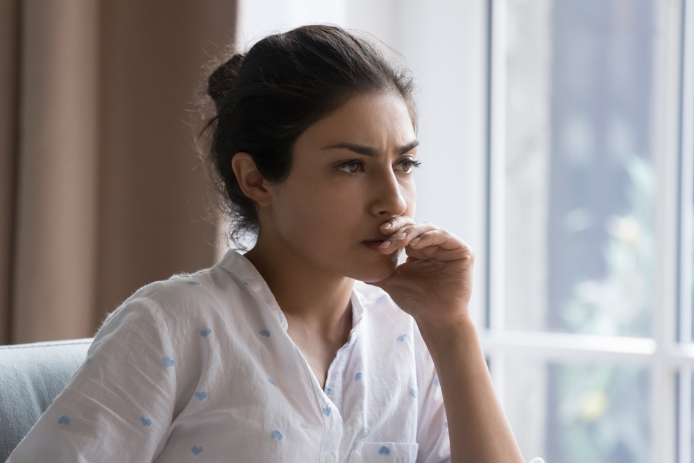 Serious thoughtful beautiful Indian woman sitting on couch indoors, touching chin, looking away, thinking over bad concerning news